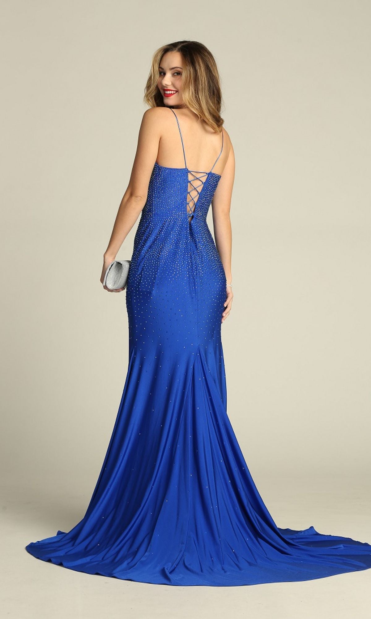 A woman faces backward and smiles, wearing the Lace-Up Long Beaded Prom Dress: Chicas YG5026 in royal blue, featuring thin straps and a lace-up back. She holds a small silver clutch.